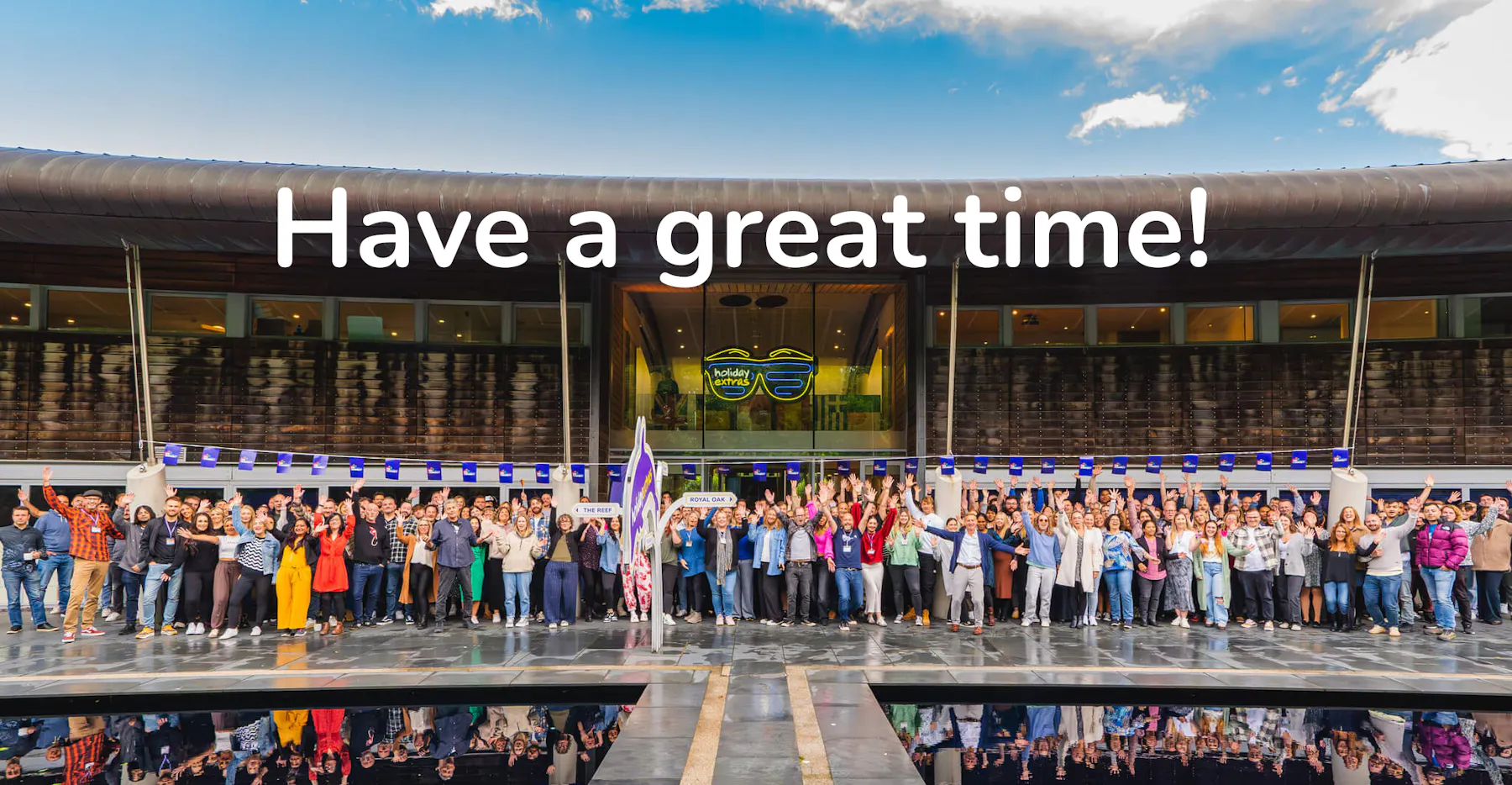 Large group of Holiday Extras staff outside a modern building with the message Have a great time above them