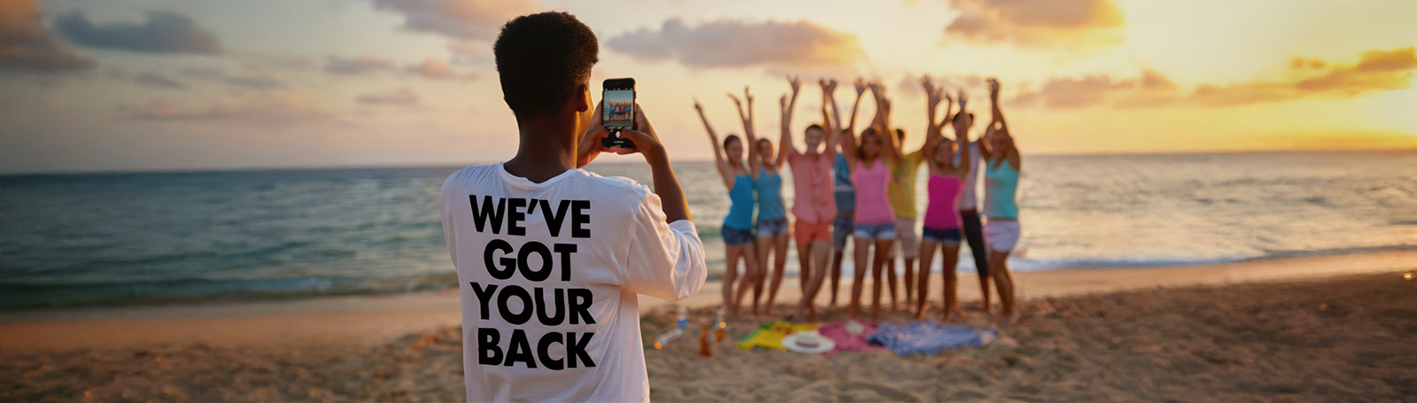 A group of friends celebrating on the beach at sunset, photographed by someone in a We've Got Your Back top